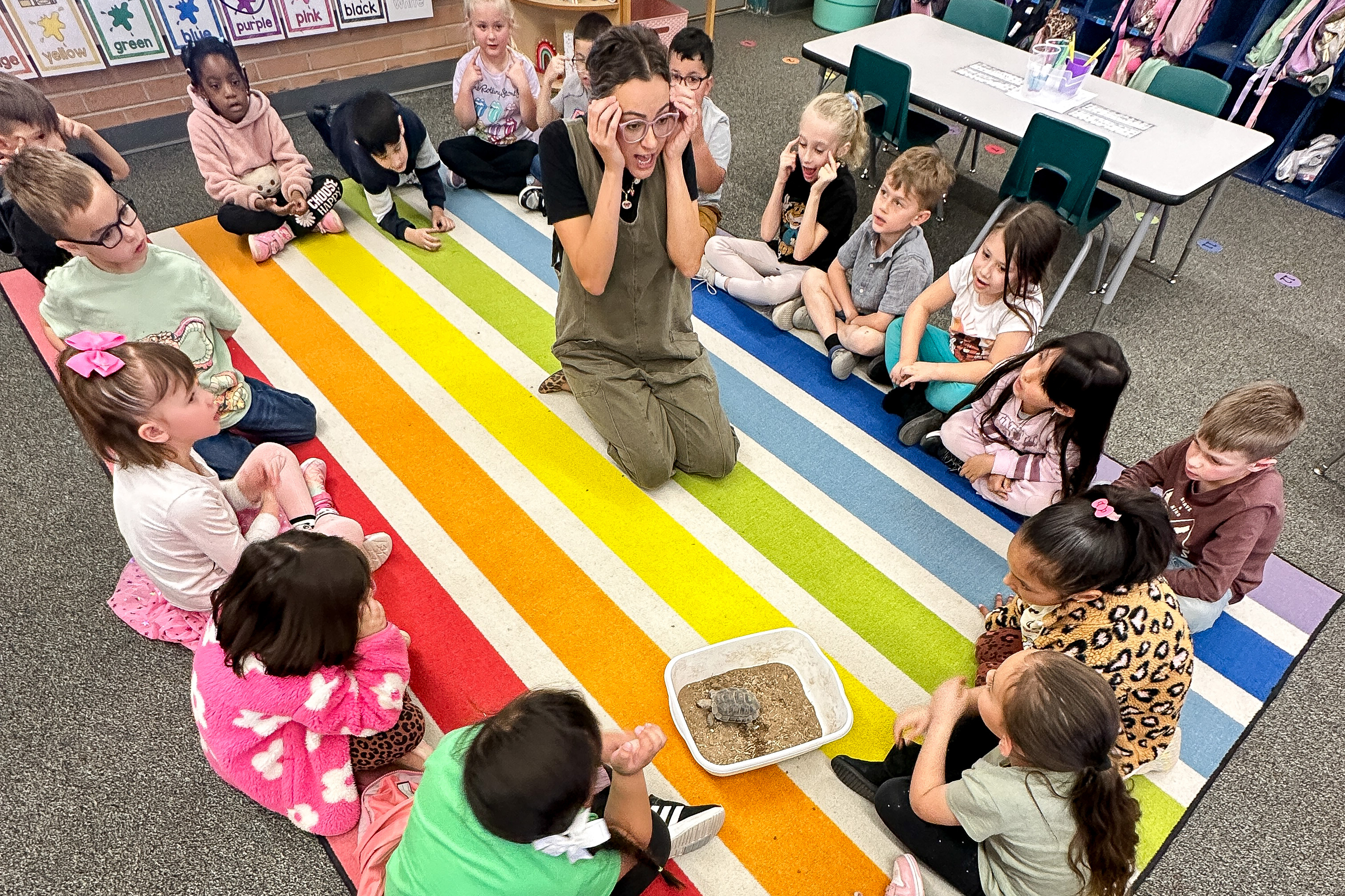 A teacher sits on a colorful rug with her students, with Tommy the desert tortoise in the corner