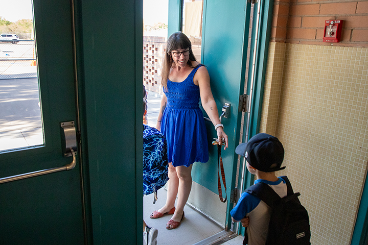 A woman in a blue dress welcomes a little boy through the school doors